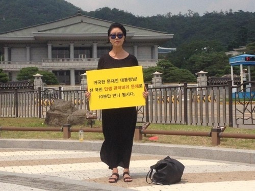 金富善手持标语站在韩国总统府青瓦台喷泉前，要求跟文在寅见面